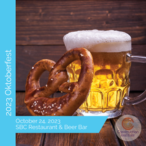 The Construction Institute's 2023 Oktoberfest. A pretzel sitting next to a glass of beer on a wooden table. The pretzel is soft and golden brown, and the beer is amber and frothy. The text on the image says 